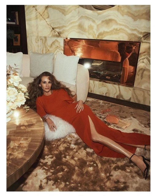 Julia Roberts lying on a shag rug in a red dress, next to a round coffee table with white roses, in an interior setting with neutral tones.
