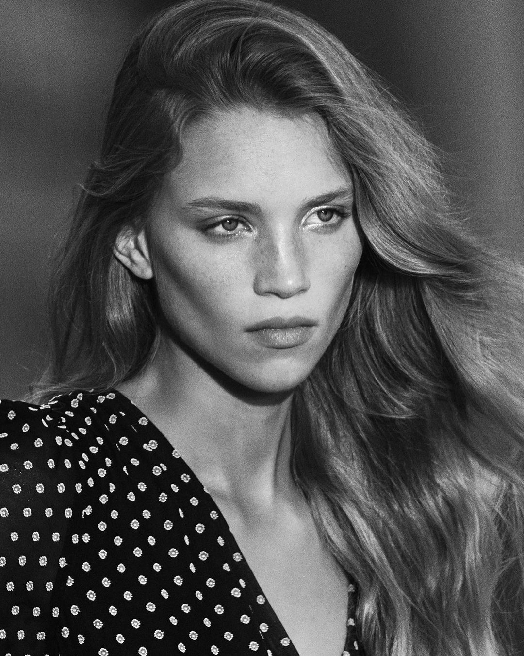 Black-and-white close-up of a young woman with long, wavy hair, wearing a polka-dot dress, in an outdoor setting with blurred background.