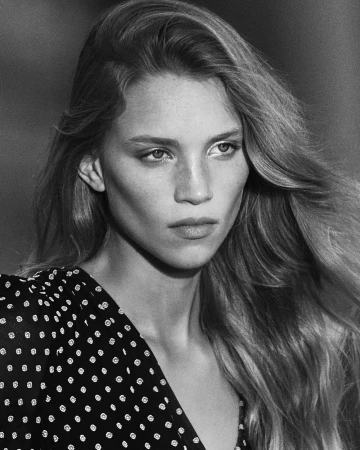 Black-and-white close-up of a young woman with long, wavy hair, wearing a polka-dot dress, in an outdoor setting with blurred background.