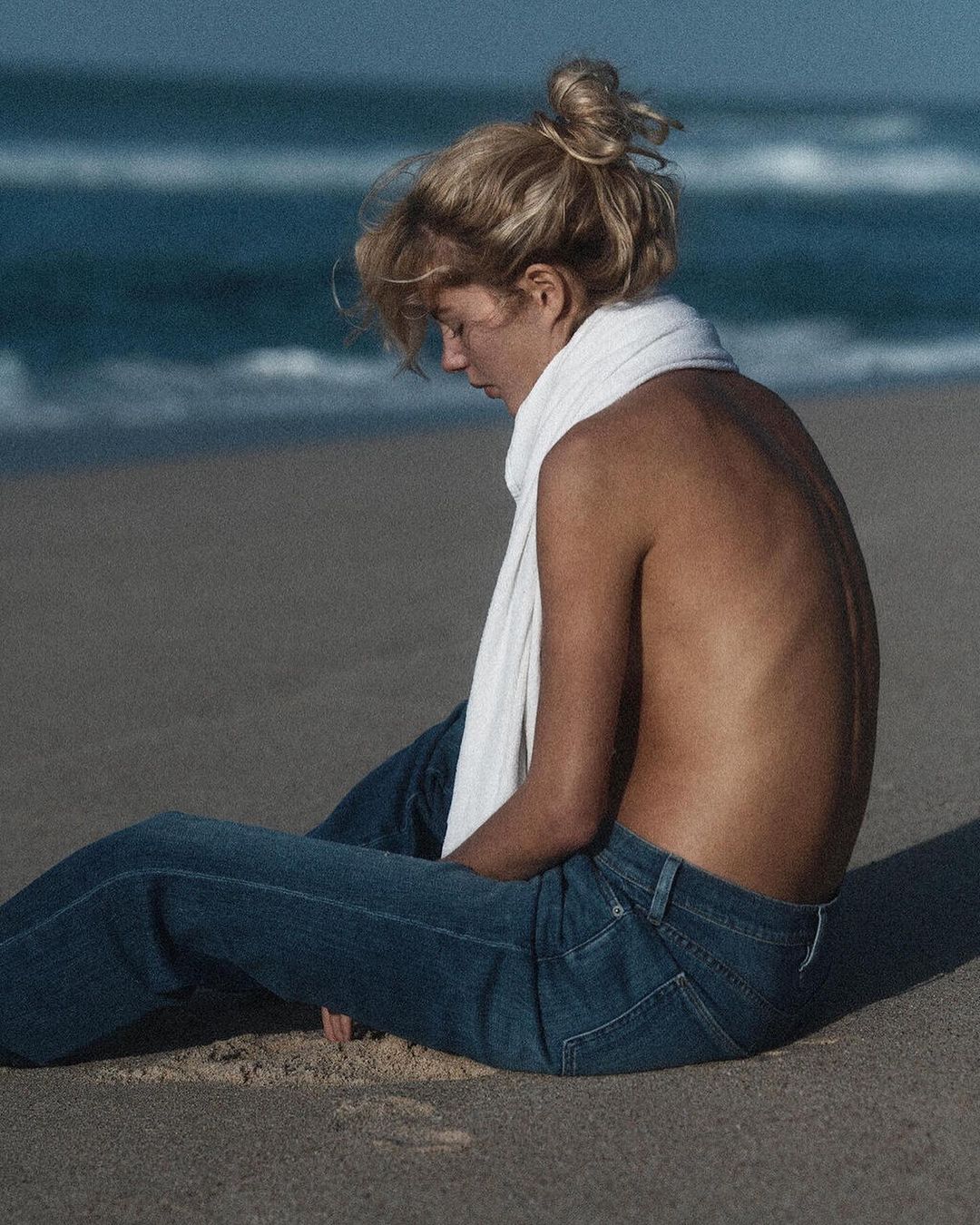 Blonde woman wearing blue jeans and a white scarf sitting on a sandy beach by the ocean, photographed by Lachlan Bailey.
