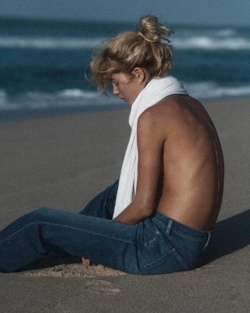 Blonde woman wearing blue jeans and a white scarf sitting on a sandy beach by the ocean, photographed by Lachlan Bailey.