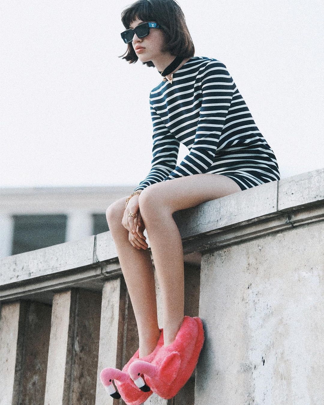 A young woman in a striped dress and flamingo slippers sits on a concrete ledge, wearing sunglasses and a black choker.
