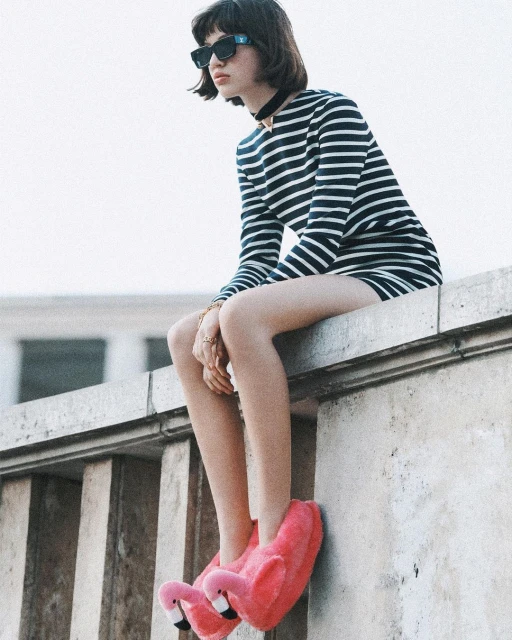 A young woman in a striped dress and flamingo slippers sits on a concrete ledge, wearing sunglasses and a black choker.