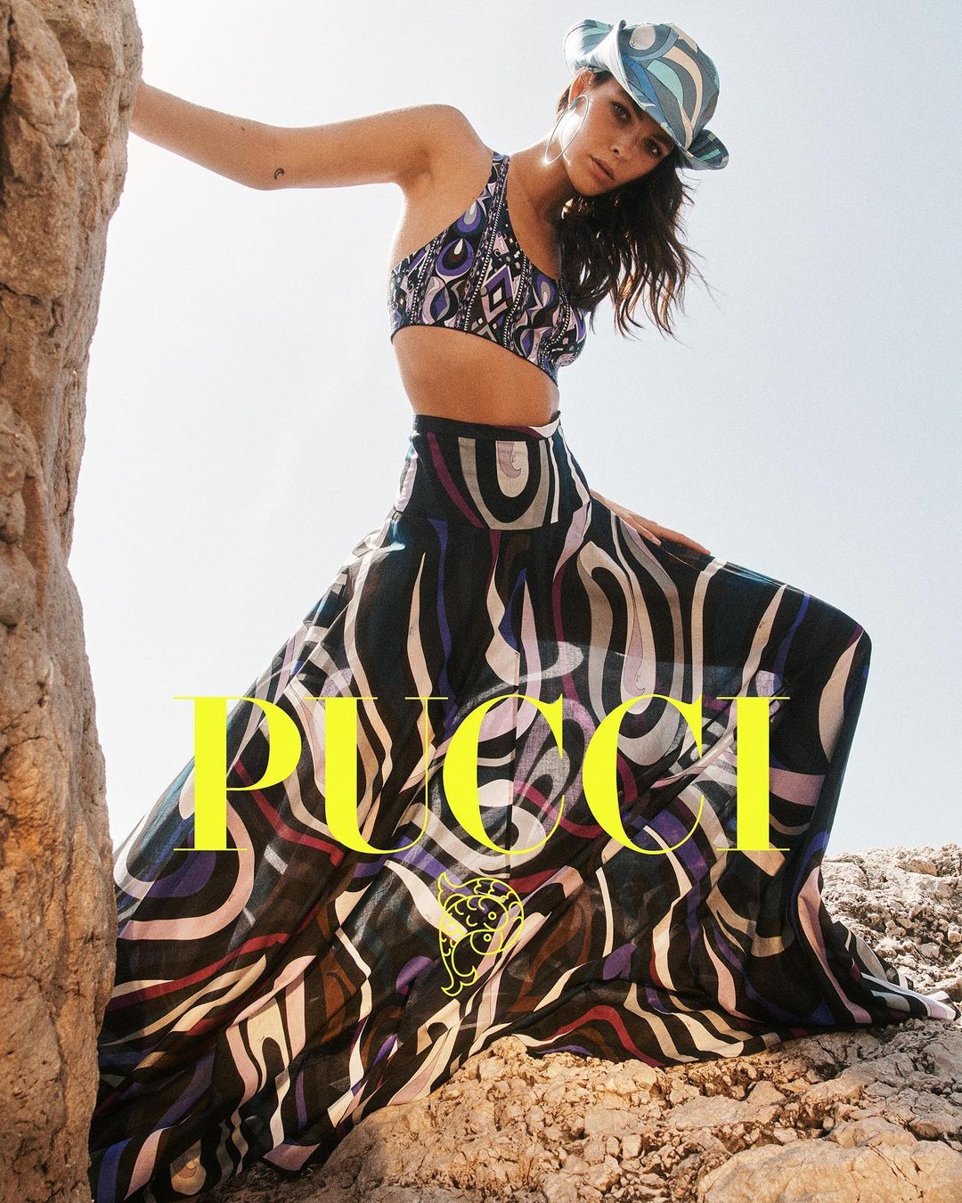 Woman poses in a patterned crop top, maxi skirt, and hat on a rocky landscape, showcasing a fashion forward style against a natural backdrop.
