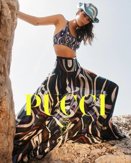 Woman poses in a patterned crop top, maxi skirt, and hat on a rocky landscape, showcasing a fashion forward style against a natural backdrop.