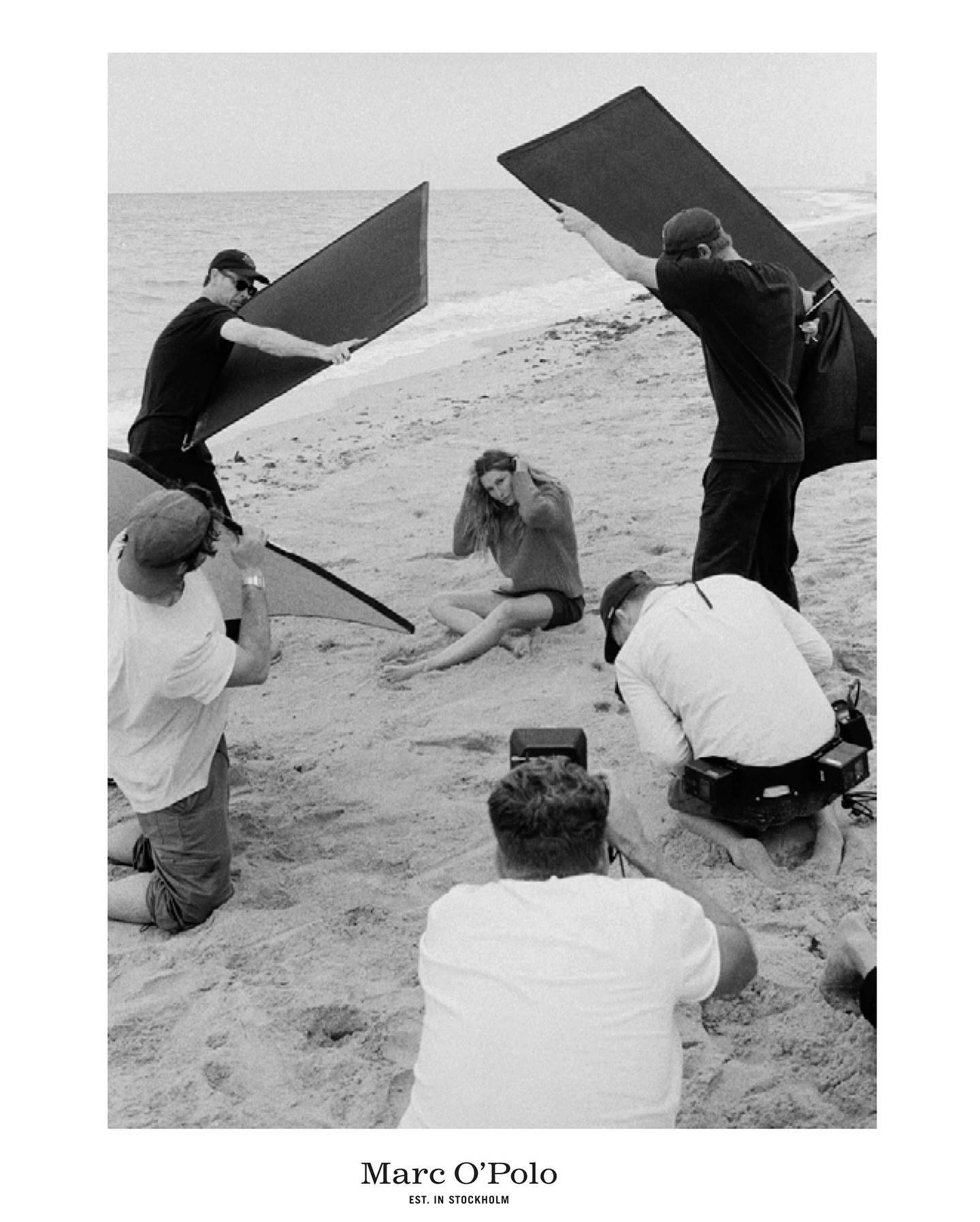 Black and white photo of a beach fashion shoot with a model sitting in the sand surrounded by photographers and reflectors.