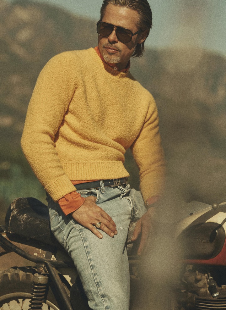 Brad Pitt poses on a red-and-white motorcycle in a sunlit outdoor scene. He wears sunglasses, a yellow sweater, and blue jeans.