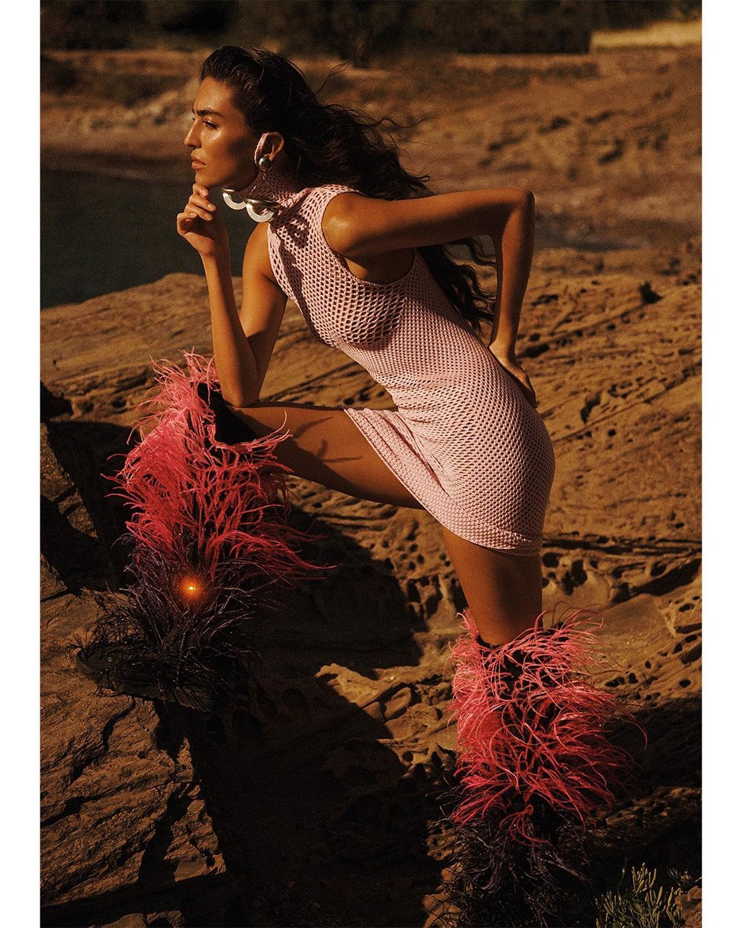 Woman in a pink knit dress and pink feather boots posing on a rocky landscape with a body of water in the background.