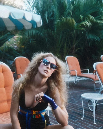 A fair-skinned adult female with long blonde hair poses on an orange lounge chair in sunglasses next to a white patio table outdoors.