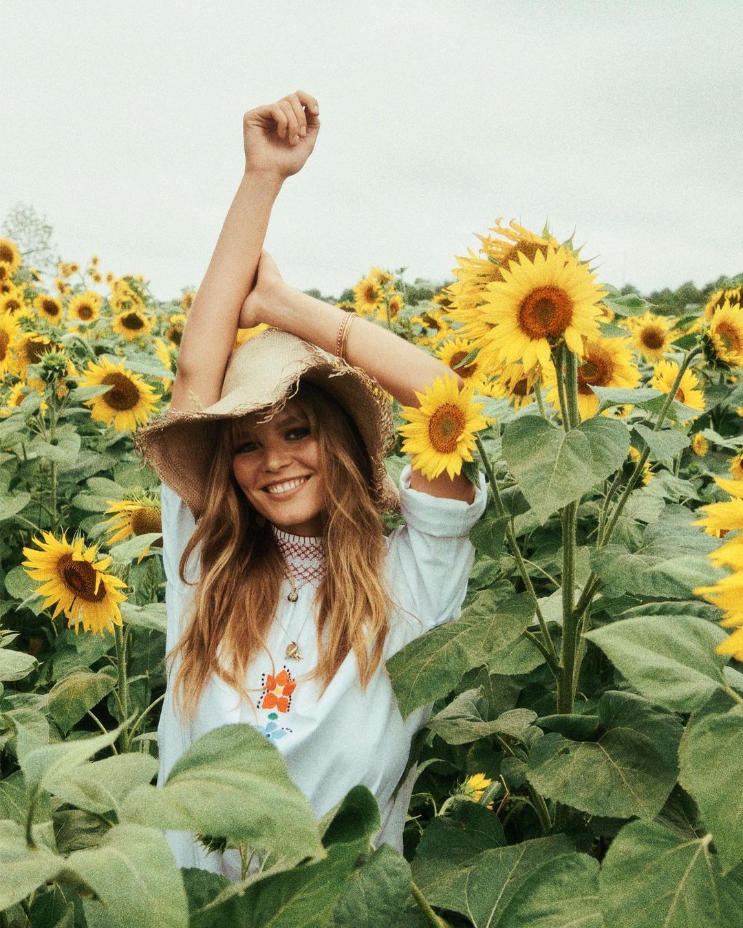Smiling woman wearing straw hat stands in a field of sunflowers with her arms up and a white cloudy sky in the background.