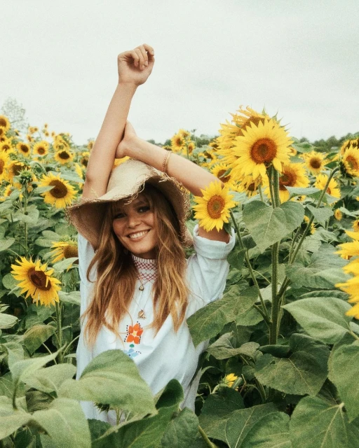 Smiling woman wearing straw hat stands in a field of sunflowers with her arms up and a white cloudy sky in the background.