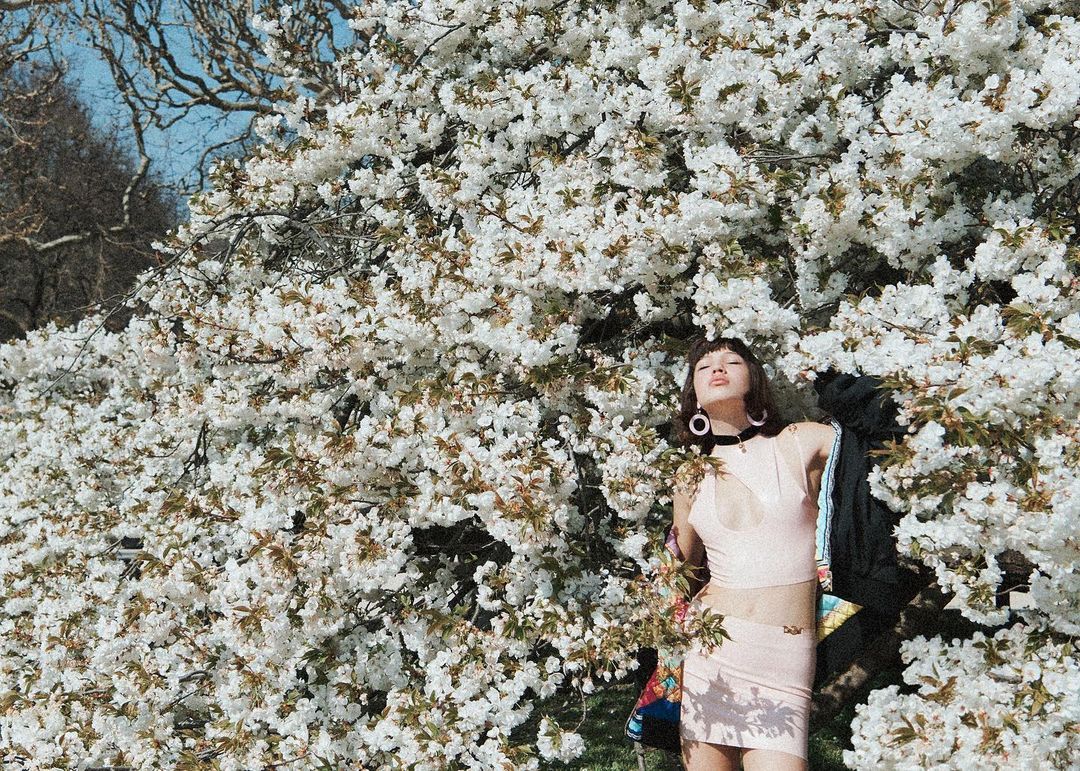 Young woman poses among blooming white tree branches in a close-up outdoor shot, wearing a pink top and skirt with a jacket on her shoulder.