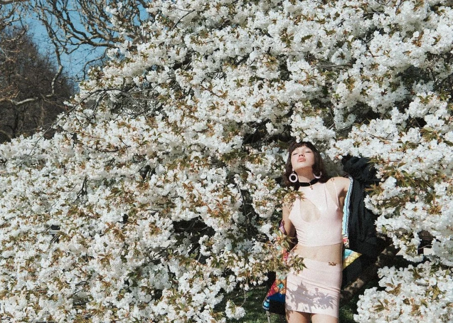 Young woman poses among blooming white tree branches in a close-up outdoor shot, wearing a pink top and skirt with a jacket on her shoulder.