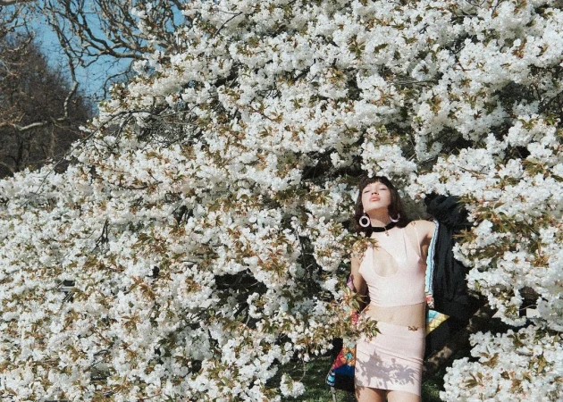 Young woman poses among blooming white tree branches in a close-up outdoor shot, wearing a pink top and skirt with a jacket on her shoulder.
