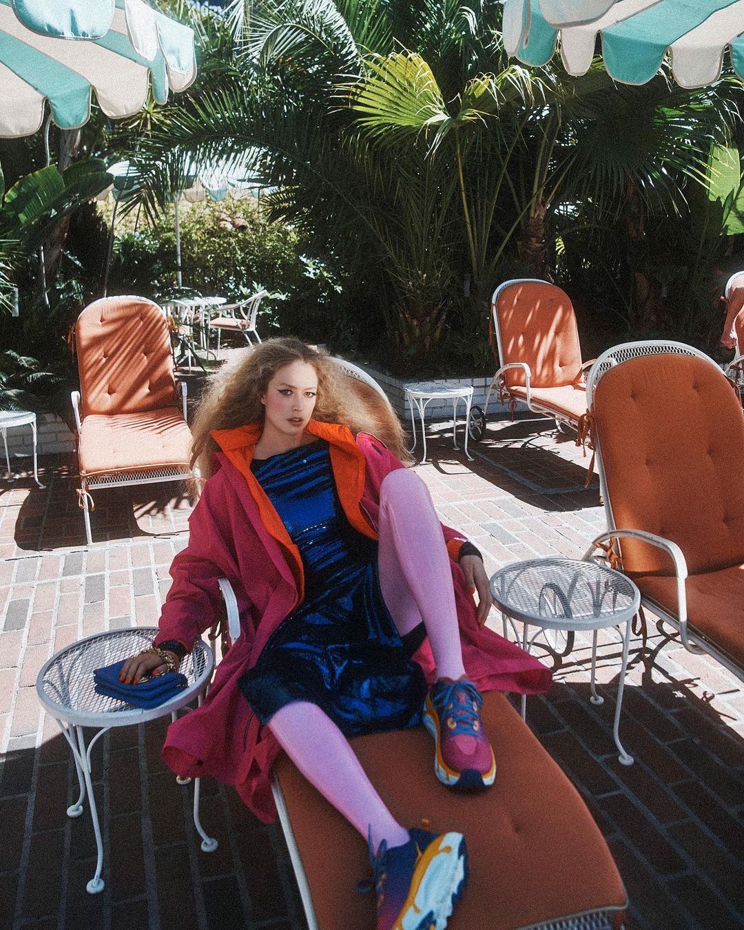 Young woman relaxes on lounge chair in bright coat, with nearby lounge seating and greenery providing a sunny outdoor ambiance.