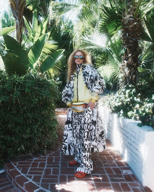 A woman with sunglasses and a patterned outfit poses on a brick path surrounded by lush greenery, creating a vibrant fashion editorial.