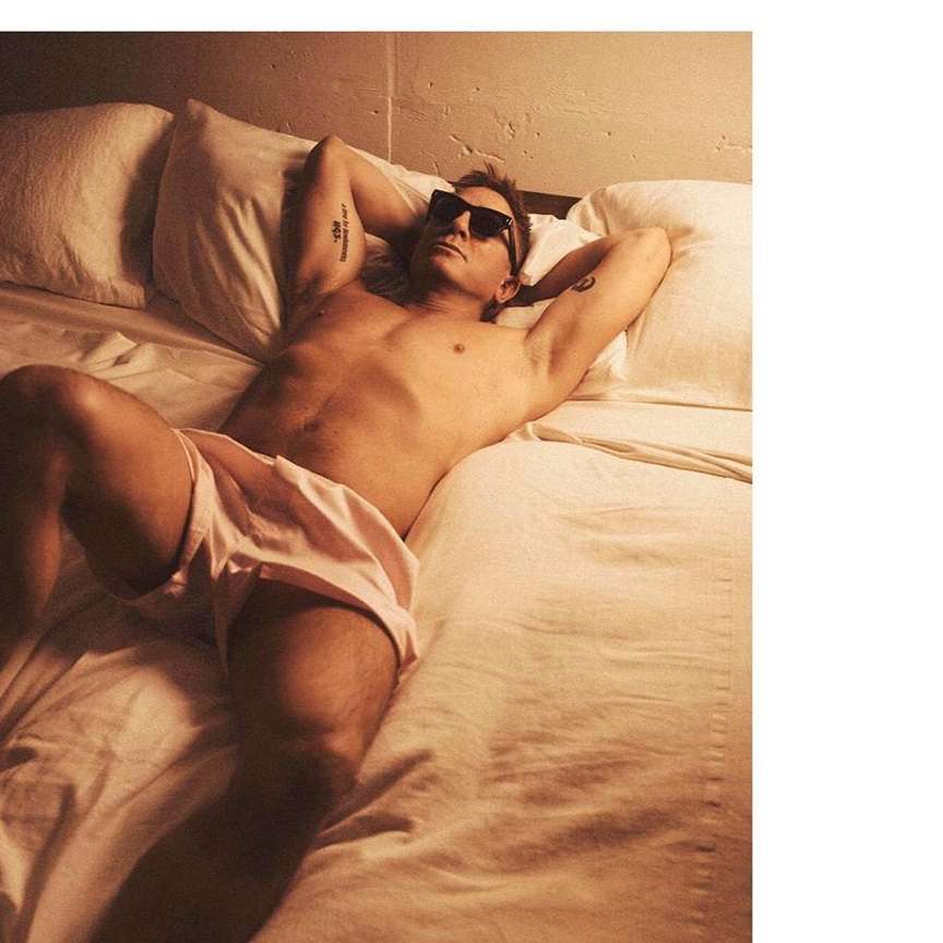 Shirtless man wearing sunglasses lying on a bed with white pillows and sheets in warm lighting