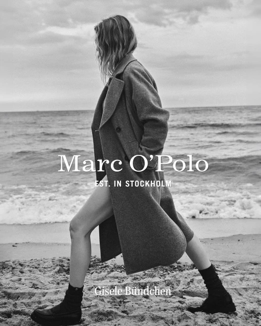 Black and white fashion shot of Gisele Bündchen in a coat walking on a sandy beach with ocean and sky backdrop in a fashion editorial.