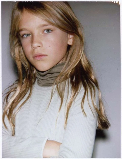 Portrait of a young girl with long blonde hair wearing a turtleneck sweater, indoors. She has blue eyes and is looking at the camera.