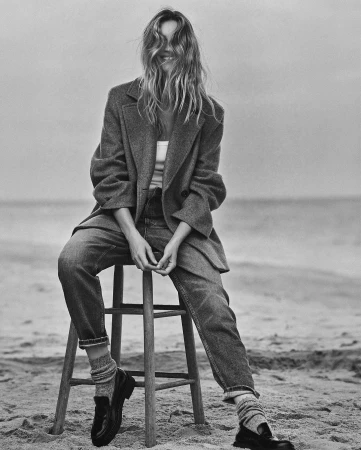 Monochrome fashion portrait of smiling woman seated on wooden stool wearing oversized wool blazer, denim trousers, loafers and ribbed socks on sandy beach