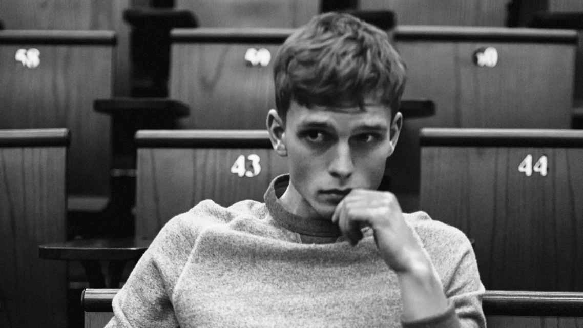 Young man with hand to face sits in a wooden auditorium. Black and white photo of a male in a crew neck sweatshirt.