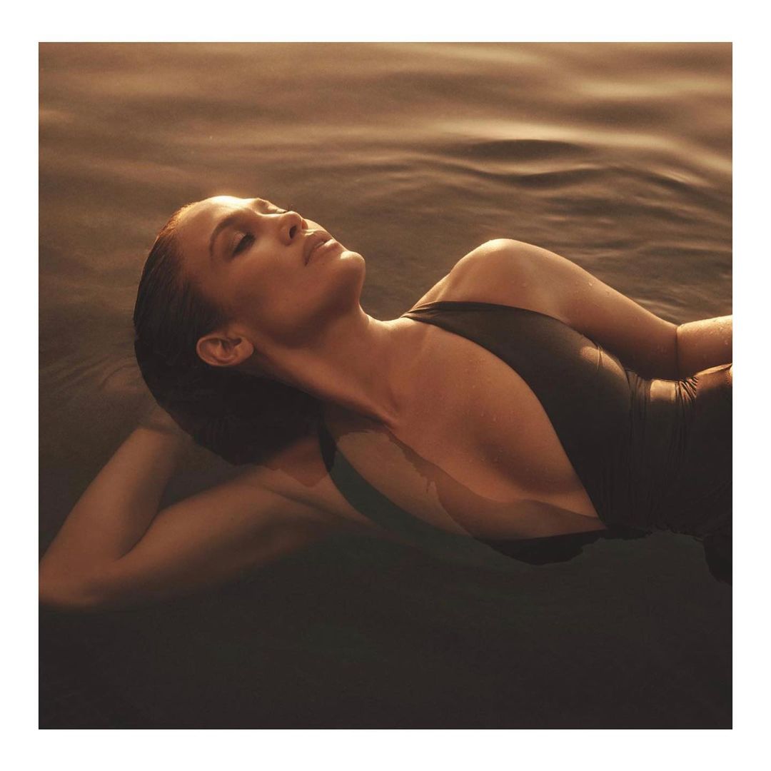 Woman with dark hair reclines in water wearing a black swimsuit, with warm, golden lighting and a serene facial expression.
