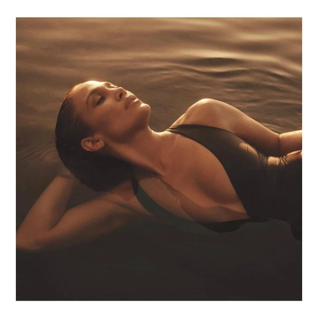 Woman with dark hair reclines in water wearing a black swimsuit, with warm, golden lighting and a serene facial expression.