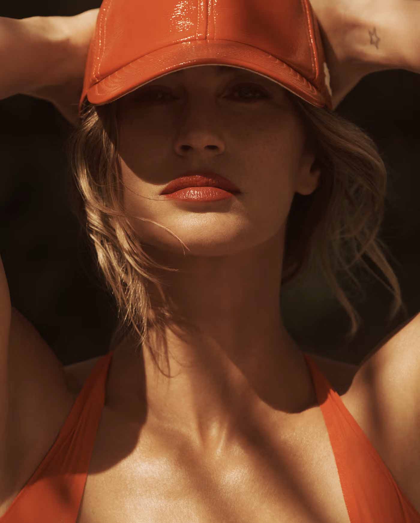 Woman wearing an orange baseball cap and matching orange top poses in a close-up shot with a neutral expression in a dark setting.