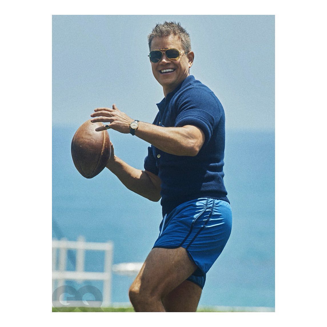 Smiling man in navy polo and blue shorts throws a football outdoors, wearing sunglasses and a watch, with a glimpse of the ocean.