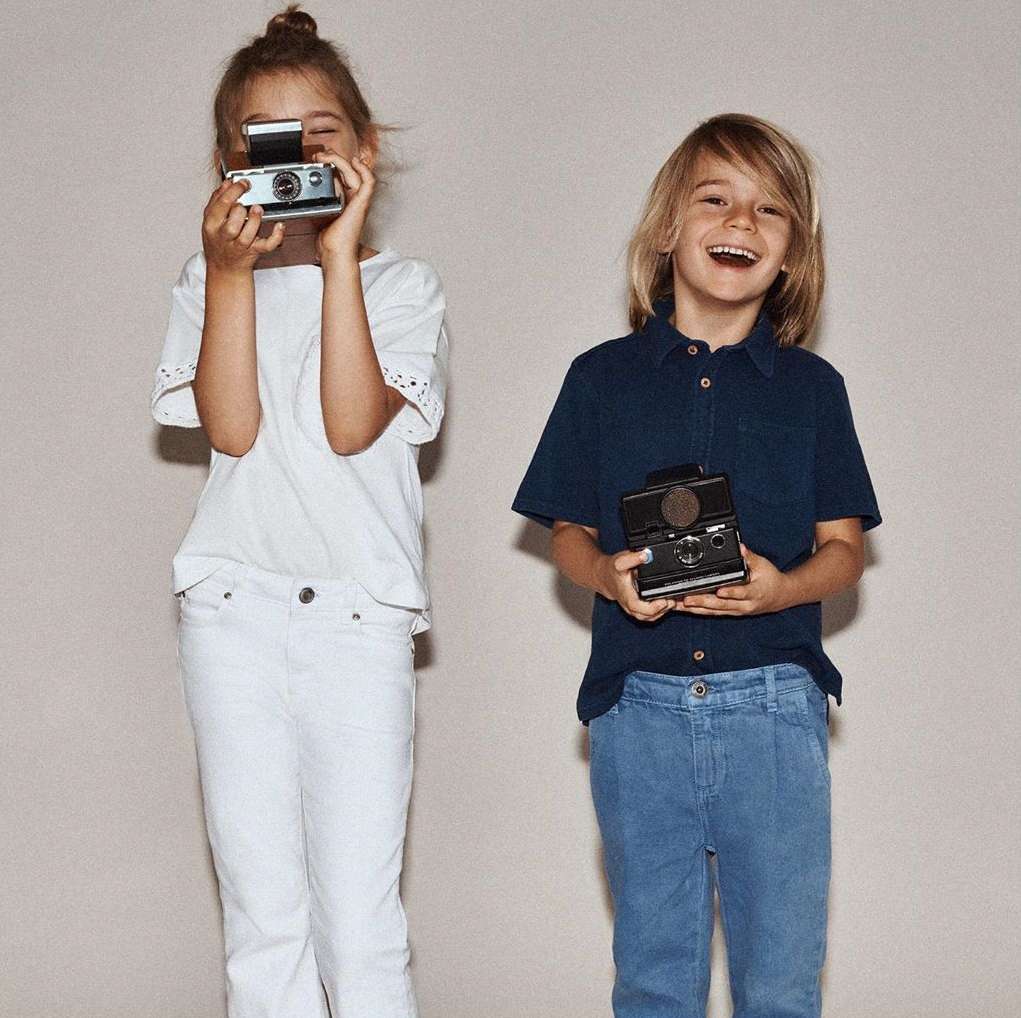 Two happy children holding vintage cameras, smiling in a studio. A girl holds a Polaroid while a boy holds a twin-lens reflex camera.