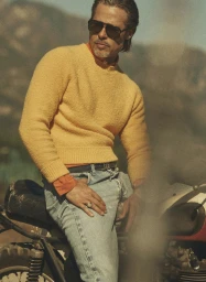 Brad Pitt wearing dark sunglasses and a textured yellow sweater while leaning on a vintage motorcycle outdoors.