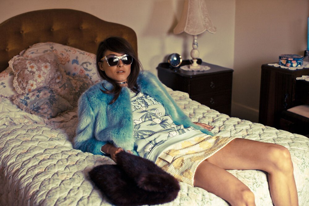 Adult female model in stylish 1960s-inspired fashion, relaxing on a bed in an indoor bedroom setting with vintage decor and soft, diffused lighting.