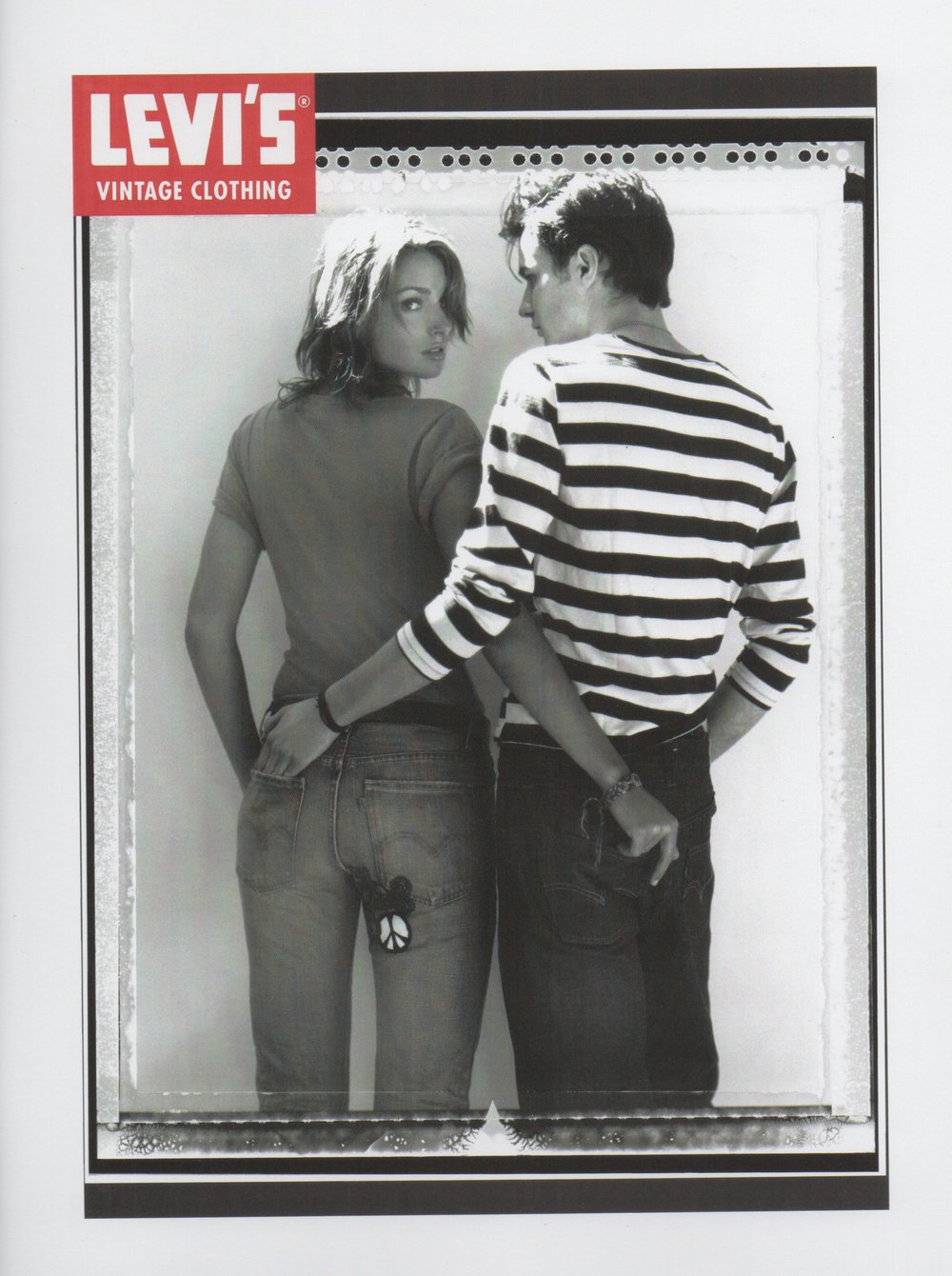 Black and white fashion shot of a young adult couple in jeans and casual tops holding hands behind their backs, posing indoors.