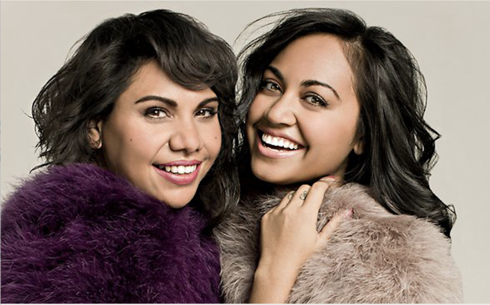 Two smiling women pose closely indoors wearing faux fur jackets; portrait shot with soft lighting and neutral background.