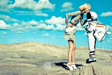Model in grey patterned jacket and white heels kissing an astronaut in a full white spacesuit on a barren rocky landscape.