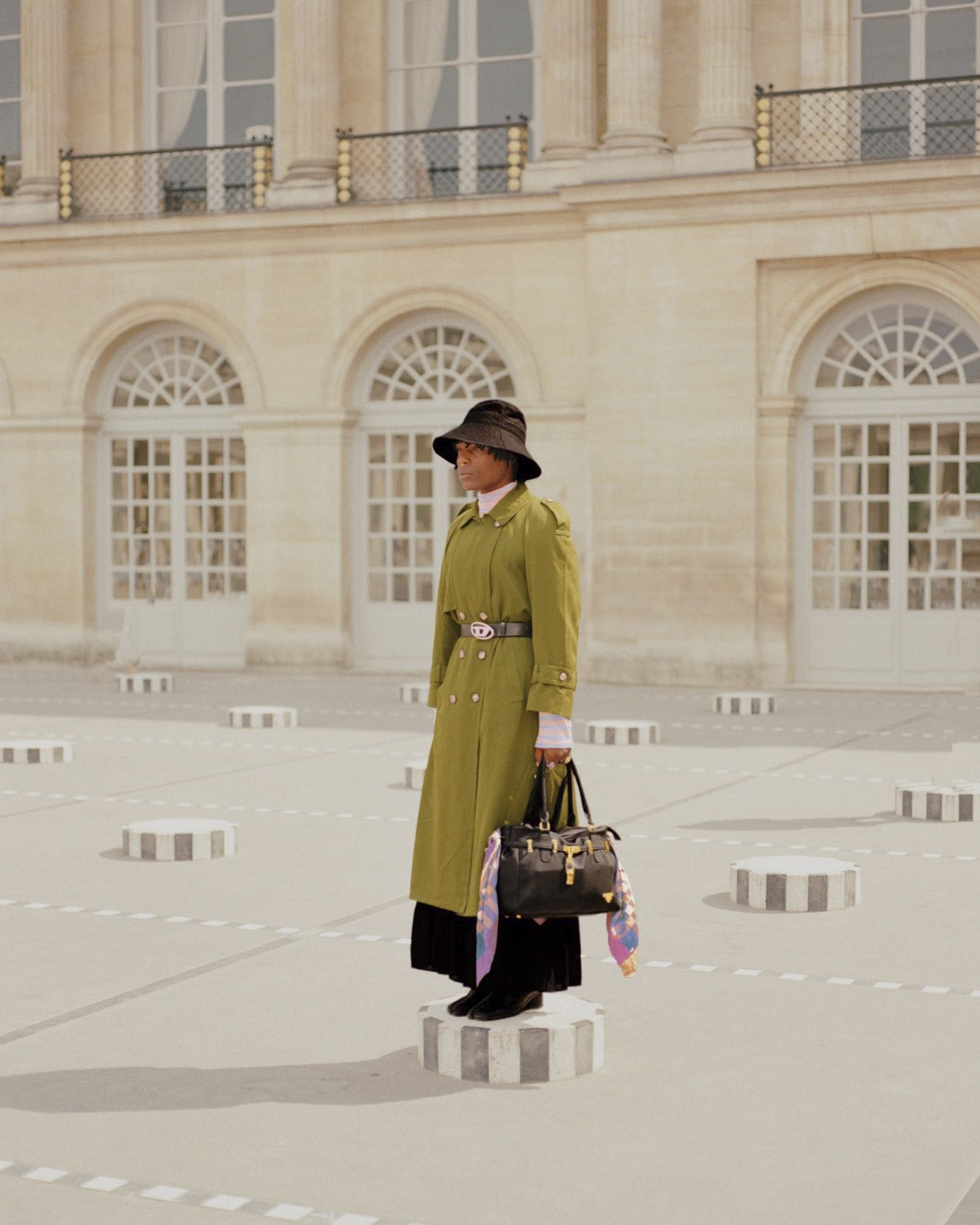 Woman in stylish olive trench coat and bucket hat posing on a pedestal in a courtyard with architectural details, carrying a black handbag.
