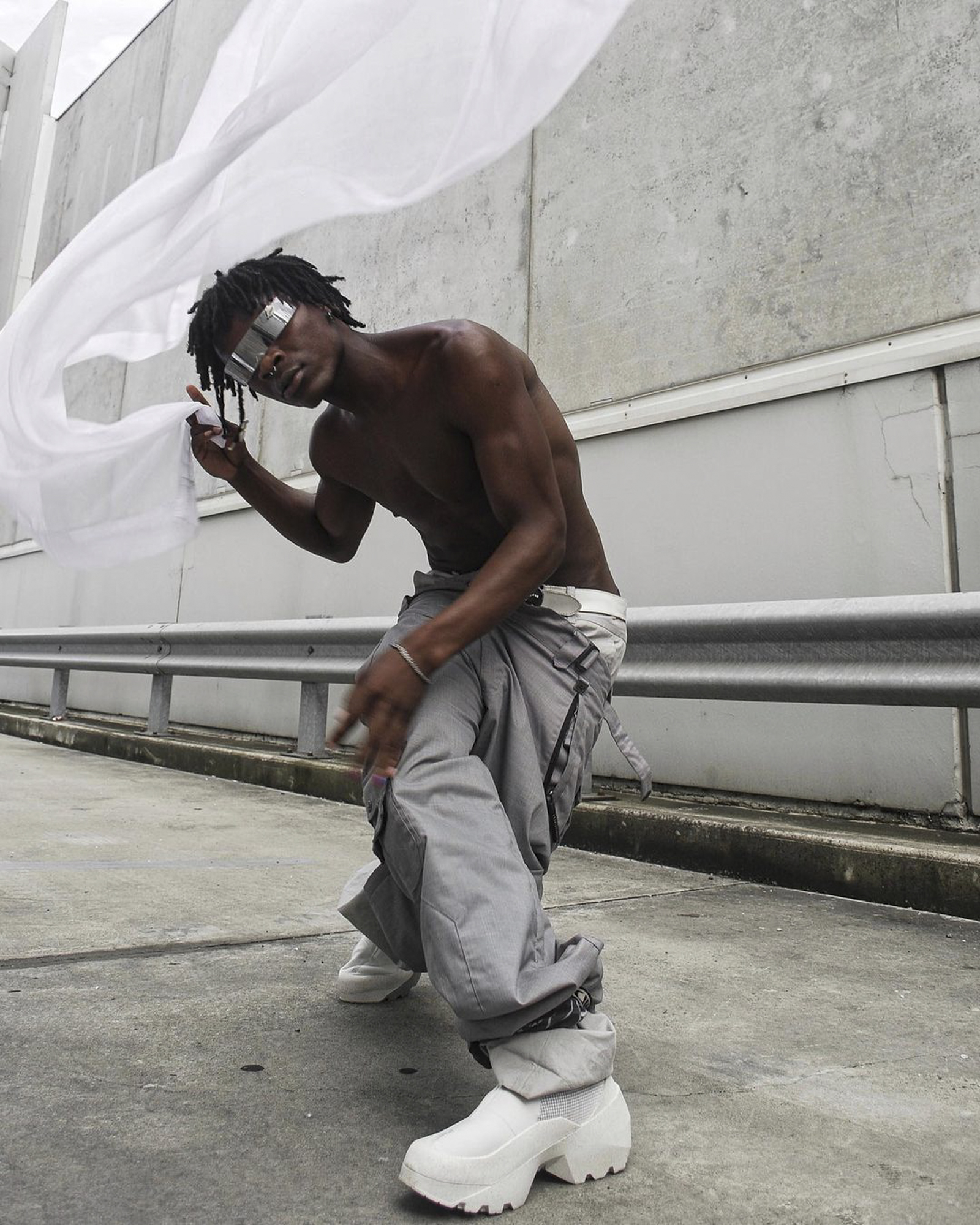 Fashion shot of an adult Black male wearing futuristic sunglasses and chunky platform boots, posing with a sheer white scarf outdoors.