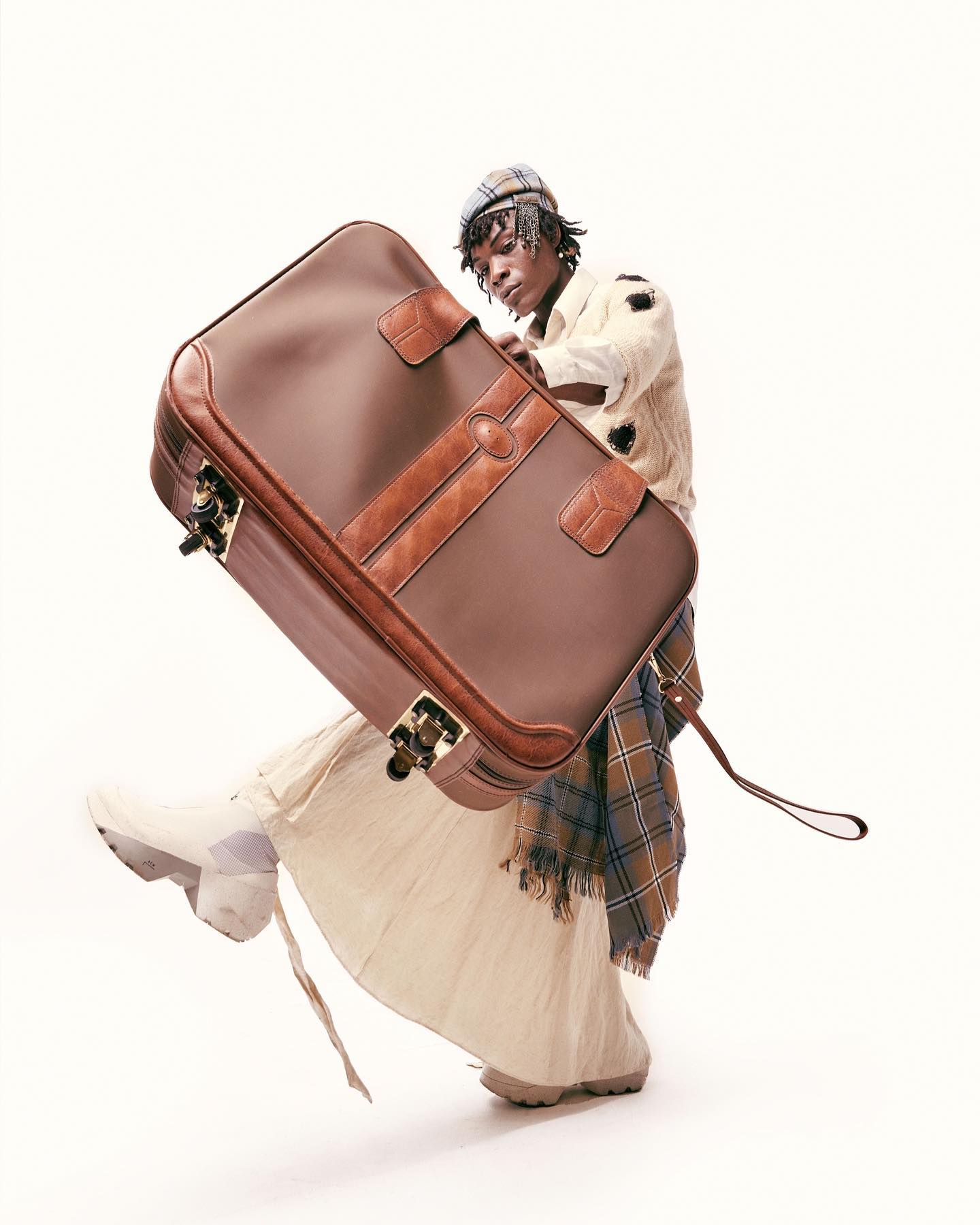 Stylish person balances a large vintage suitcase with a plaid scarf. They wear a matching plaid hat and cream-colored jacket in a studio.
