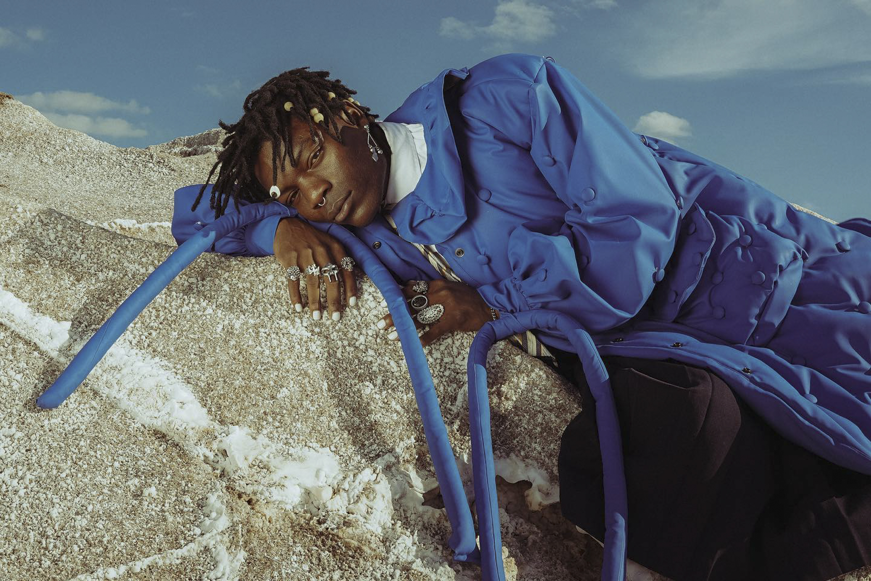 Portrait of Black male model with dreadlocks reclining on rock wearing a royal blue puffer coat in an outdoor, sunny, daytime setting.