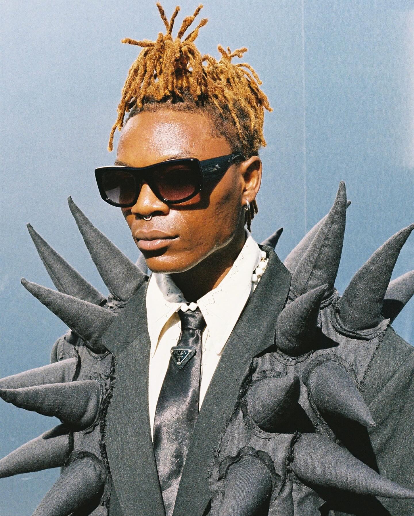 Portrait of a Black male model with dreadlocks wearing sunglasses, a jacket with spikes, and Prada accessories against a grey backdrop.
