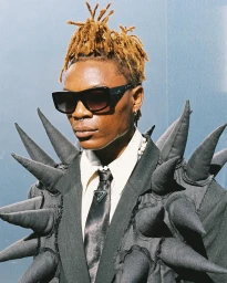 Close-up portrait of a Black man with dreadlocks wearing dark sunglasses, a white shirt, a black tie, and a spike-covered jacket by Bernad Ngendakumana.