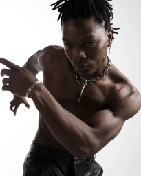 Shirtless black man wearing layered silver chains, septum ring, and dark leather trousers against a white backdrop.