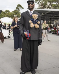 Man wearing charcoal pinstripe suit, wide black trousers, cap, and sunglasses with plush toy pinned to jacket by Bernad Ngendakumana