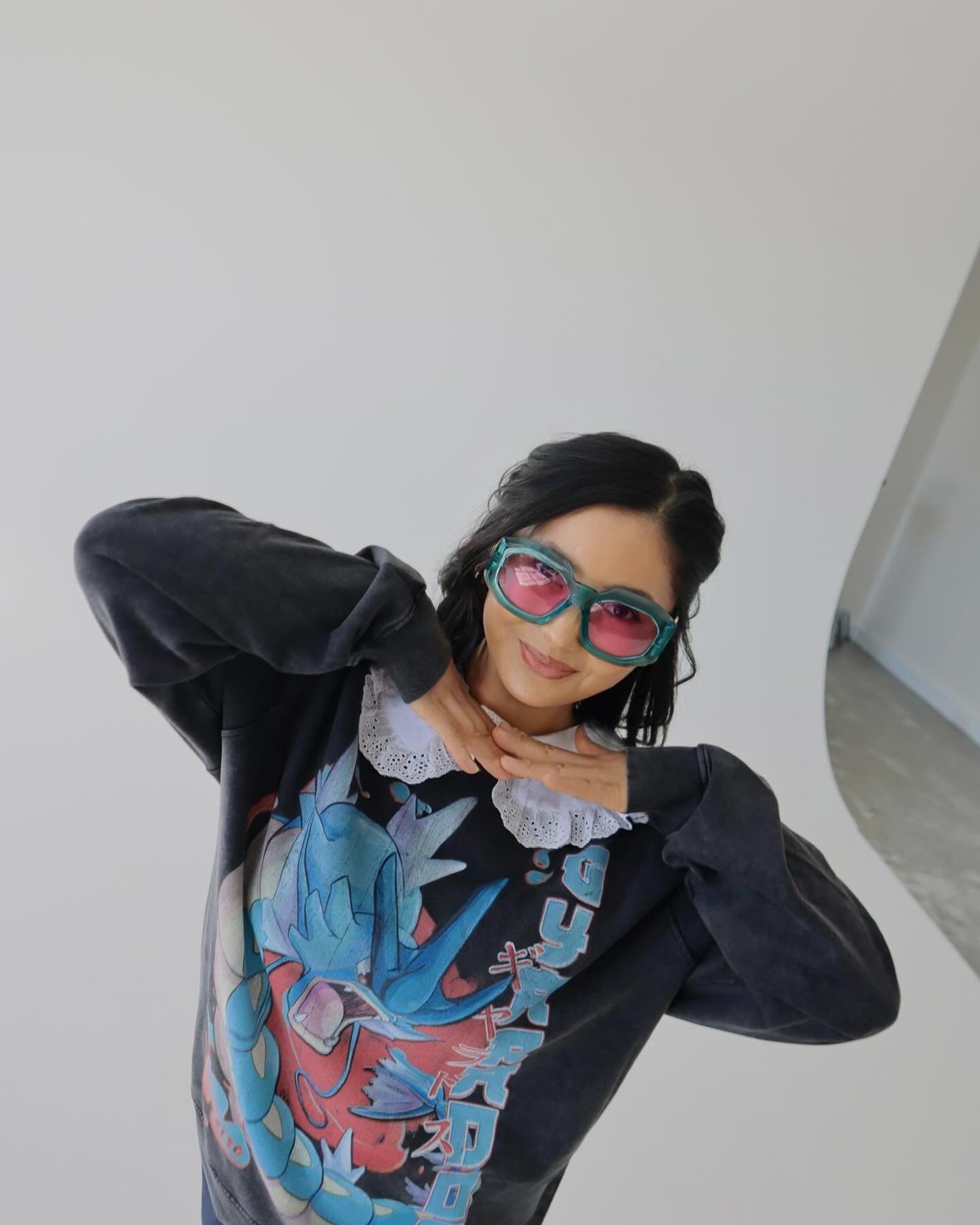 An adult female poses in a studio setting, wearing a sweatshirt with a large graphic, complemented by bright sunglasses.