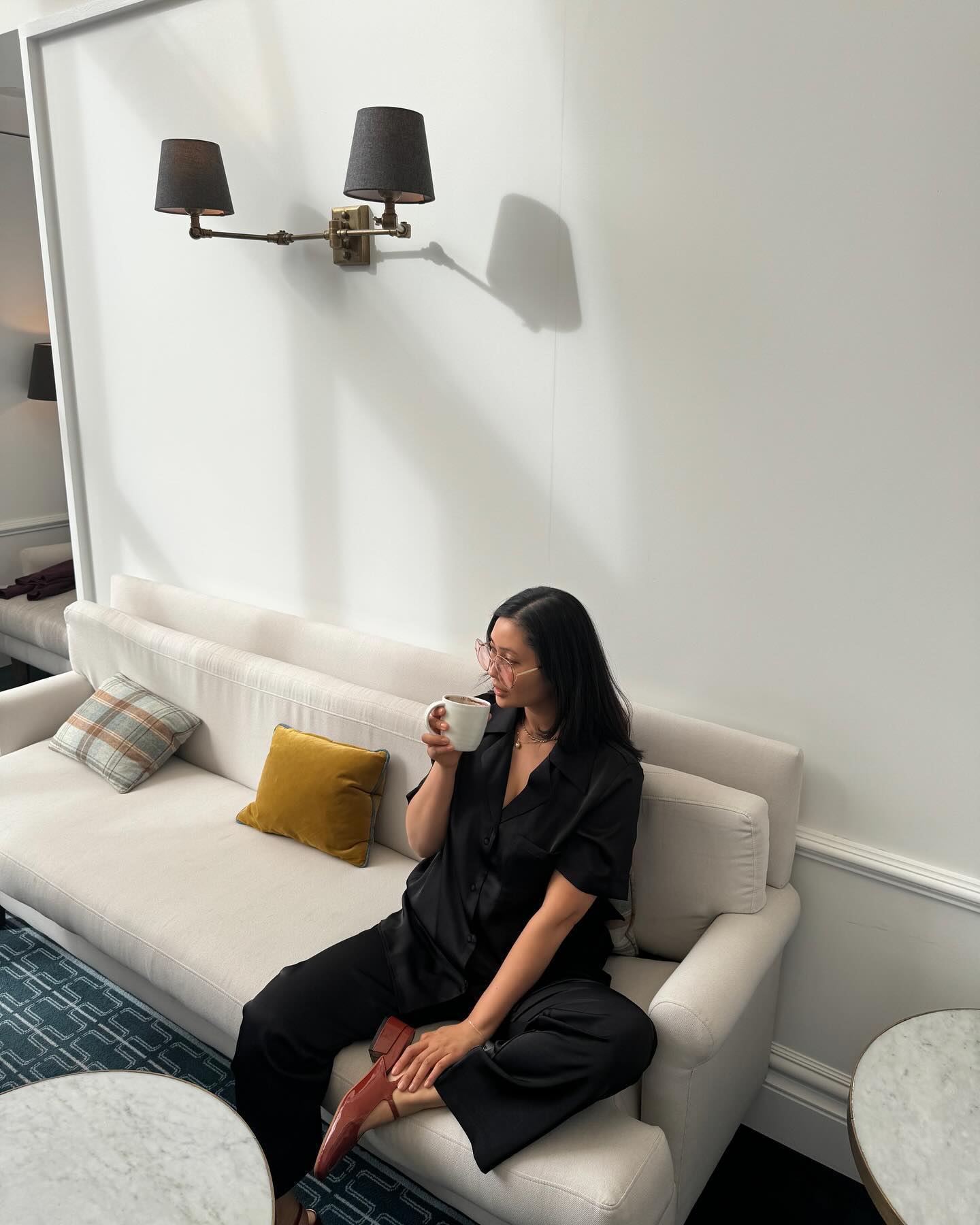 A woman in black loungewear sits on a cream sofa in a bright room, holding a mug, with a brass sconce and pillows visible.