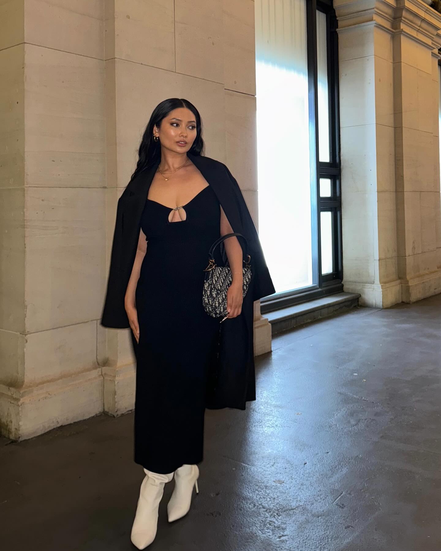Asian woman in a black dress and white boots poses indoors with a black coat draped over her shoulders, holding a patterned handbag.