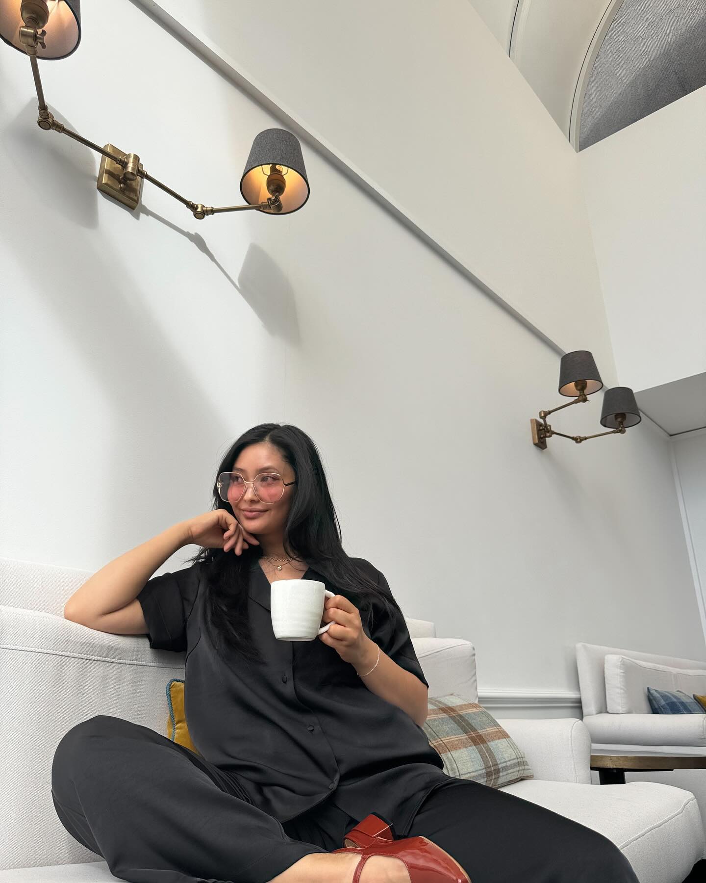 Woman in black loungewear sits on white sofa with coffee, wearing pink glasses, with retro wall lamps in a brightly lit room.