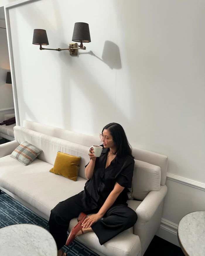 Woman in black silk pajama set seated on a cream sofa drinking from a mug inside a brightly lit Melbourne interior space.