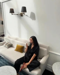 Woman in black silk pajama set seated on a cream sofa drinking from a mug inside a brightly lit Melbourne interior space.