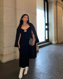 Woman in a black dress holding a patterned handbag and wearing white boots standing outdoors